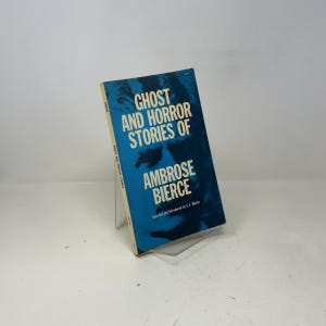 Puede incluir: Un libro de bolsillo vintage titulado "Ghost and Horror Stories of Ambrose Bierce". La portada es un degradado de azul y blanco con texto blanco en negrita. El libro está apoyado en un soporte transparente.