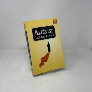 Op de afbeelding: Een hardcover boek getiteld "The Alienist" van Caleb Carr. Het boek heeft een gele omslag met een zwart-wit illustratie van een persoon en een schaduw. De woorden "The Alienist" staan in een zwart kader.