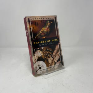 Puede incluir: Un libro titulado "Empires of Time" de Anthony Aveni. La portada presenta una ilustración de un pájaro y esferas de reloj. El libro se exhibe en un soporte de plástico transparente.