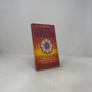 Puede incluir: Un libro de bolsillo titulado Creative Visualization de Shakti Gawain con una portada en degradado rojo y amarillo. La portada presenta un diseño geométrico de mandala en forma de estrella en el centro, con el texto Over 2 Million Copies Sold arriba.