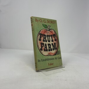 May include: A vintage book titled "The Fruit Farm" by O.G. Dorey. The cover features a large apple illustration with the title in bold letters. The book is green with red and brown accents and is displayed on a clear stand.