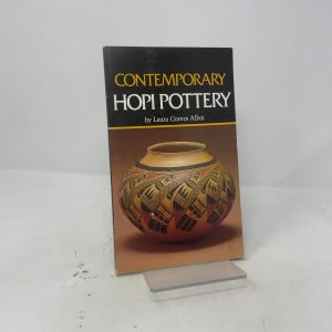 May include: A softcover book titled Contemporary Hopi Pottery by Laura Graves Allen. The cover features a black top section with yellow and white text, and a lower section displaying a round, tan-colored pottery vessel with black geometric patterns.