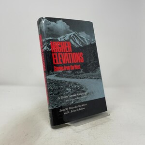 Puede incluir: Un libro de tapa dura titulado Higher Elevations: Stories from the West, editado por Alexander Blackburn y C. Kenneth Pellow. La portada presenta un paisaje en blanco y negro de una montaña y un camino sinuoso, con el título en texto rojo.