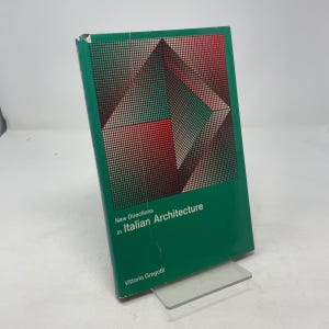 以下が含まれることがあります： ヴィットリオ・グレゴッティ著「New Directions in Italian Architecture」というタイトルの緑色のハードカバー本。表紙は赤、緑、黒の幾何学模様です。本は透明なプラスチック製のスタンドに立てかけられています。