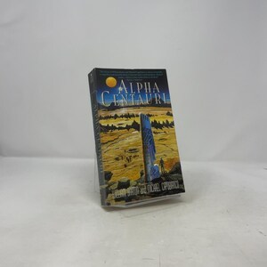 May include: A hardcover book titled "Alpha Centauri" with a cover depicting a landscape with a blue structure and a yellow sun. The author's names are at the bottom. The book is set against a white background.