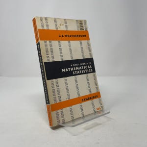 Puede incluir: Un libro antiguo titulado "A First Course in Mathematical Statistics" de C.E. Weatherburn. El libro tiene una cubierta blanca con detalles en naranja y texto negro. El libro está apoyado en un soporte transparente.