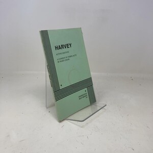 May include: A light green book titled "HARVEY" with black text, including "ACTING EDITION" and "A COMEDY IN THREE ACTS BY MARY CHASE." The book has black horizontal stripes and stars. It is displayed on a clear stand.