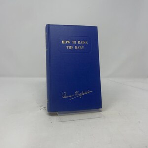 May include: A hardcover book titled "HOW TO RAISE THE BABY" in gold lettering on a blue cover. The author's signature is at the bottom. The book is standing upright on a clear stand.