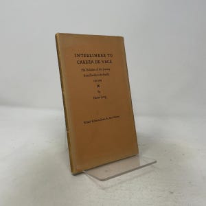 May include: A tan hardback book titled Interlinear to Cabeza de Vaca, His Relation of the Journey from Florida to the Pacific 1528-1536 by Haniel Long, published by Writers' Editions, Santa Fe, New Mexico, standing upright on a clear plastic display stand.