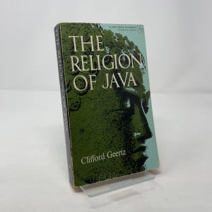 Peut inclure: Un livre de poche intitulé "The Religion of Java" par Clifford Geertz. La couverture présente une illustration verte d'un visage et le titre en blanc. Le livre est présenté sur un présentoir en plastique transparent.