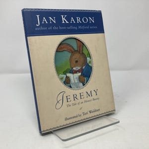 May include: A hardcover book titled "Jeremy" by Jan Karon, featuring an illustration of a bunny reading. The book has a blue spine and a cream-colored cover with the title and author's name prominently displayed.