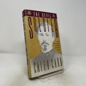 Op de afbeelding: Een hardcover boek getiteld The Devil Soldier van Caleb Carr. De kaft bevat een sepia-kleurig portret van een man met een snor tegen een getextureerde gouden achtergrond. De titel is gedrukt in grote donkerrode en zwarte letters.