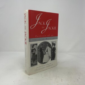 May include: A hardcover book titled Jack and Jackie: Portrait of an American Marriage by Christopher Andersen. The cover features a red top section with white text and a bottom section with three black and white photographs of a couple.