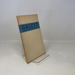 May include: A vintage book titled "GALCIT The First Twenty-Five Years." The book has a light brown cover with a blue band across the top. The text is in white and the book is displayed on a clear stand.