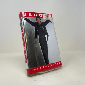 May include: A VHS tape titled Jagger Unauthorized featuring a photograph of a person in a black shirt and black trousers with arms raised. The cover has a red border at the top and bottom with white text. A small white price sticker is on the top.