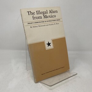 May include: A book titled "The Illegal Alien from Mexico" with a tan and white cover. The title is in black text, with the subtitle "Policy Choices for an Intractable Issue." A black star is in a white square on the cover.