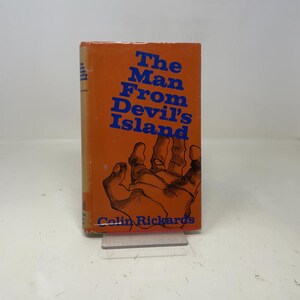 May include: An orange book titled "The Man From Devil's Island" with blue text and a hand illustration. The author's name, Colin Richards, is printed at the bottom. The book is displayed on a clear plastic stand.