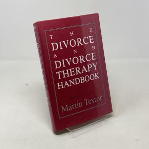 May include: A hardcover book titled "The Divorce and Divorce Therapy Handbook" in a red cover. The title is in white, with the editor's name, Martin Textor, at the bottom. The book is displayed on a clear stand.