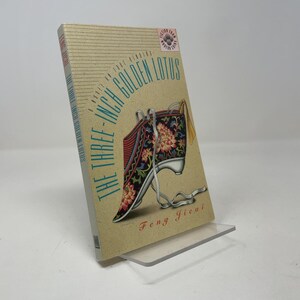 May include: A paperback book titled The Three-Inch Golden Lotus by Feng Jicui. The cover features a tan background with horizontal lines and a colorful illustration of a lotus-patterned shoe with white ribbons. A small circular sticker is at the top.