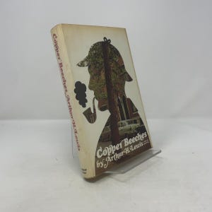 May include: A vintage book titled "Copper Beeches" by Arthur H. Lewis. The cover features a silhouette of a man in a hat smoking a pipe, with a background of trees and buildings. The book is displayed on a clear stand.