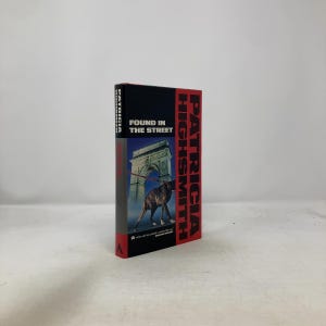 May include: A hardcover book titled "Found in the Street" by Patricia Highsmith. The cover features a dog walking under an archway, with the title and author's name in bold, red and white text against a black background.