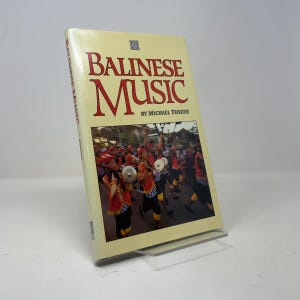 以下が含まれることがあります： マイケル・テンザー著『Balinese Music』のハードカバー本。クリーム色の表紙で、表紙には赤と黄色の伝統的な衣装を身にまとい、屋外のパレードでシンバルを持っている演奏者たちの写真が印刷されています。