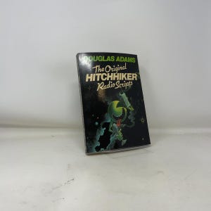 May include: A hardcover book titled "The Original Hitchhiker Radio Scripts" by Douglas Adams. The book cover is black with green and white text and a green alien illustration. The book is slightly angled.