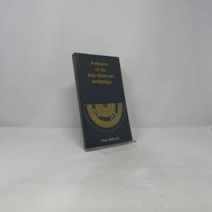 Peut inclure: Un livre à couverture rigide intitulé "Prehistory of the Indo-Malaysian Archipelago" par Peter Bellwood. Le livre a une couverture bleu foncé avec des lettres dorées et un motif décoratif doré. Le livre est posé sur un support transparent.