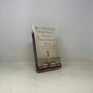 Pode incluir: Um livro de capa dura intitulado Dogs That Know When Their Owners Are Coming Home and Other Unexplained Powers of Animals de Rupert Sheldrake. A capa mostra um cão branco sentado num chão de azulejos em frente a uma porta fechada.
