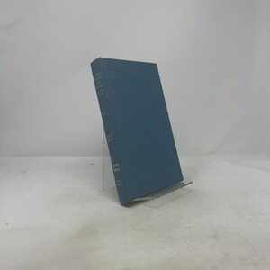 Könnte beinhalten: Ein blaugrünes Buch mit Text auf dem Buchrücken, das auf einem transparenten Ständer steht. Der Einband des Buches ist einfarbig blaugrün. Der Buchrücken enthält weißen Text, einschließlich Titel und Autorenname. Das Buch scheint eine gebundene Ausgabe zu sein.