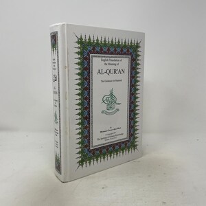 May include: A white hardback book titled English Translation of the Meaning of AL-QUR'AN The Guidance for Mankind. The cover features a decorative green, blue, and red border with intricate patterns and a central green calligraphic emblem.