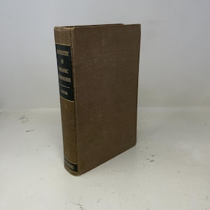 Op de afbeelding: Een hardcover boek getiteld "Chemistry of Organic Compounds" door Karrer en Heller. Het boek heeft een bruine kaft en rug met zwarte tekst. Het boek staat rechtop op een wit oppervlak.
