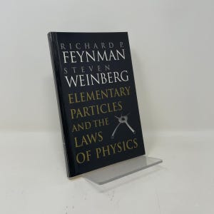 May include: A black book titled Elementary Particles and the Laws of Physics by Richard P. Feynman and Steven Weinberg. The cover features white and gold text with a small grey diagram of a particle interaction on a dark background.