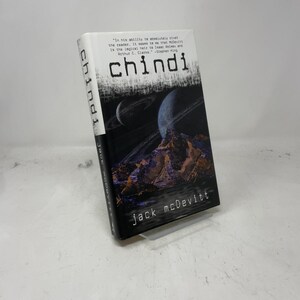 Può includere: Un libro tascabile intitolato "Chindi" di Jack McDevitt. La copertina presenta una scena spaziale scura con pianeti e una catena montuosa. Il titolo è in grandi lettere bianche.