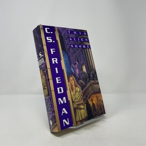 May include: A hardcover book titled "This Alien Shore" by C.S. Friedman. The cover features a woman in a gold dress, a dark building, and the title in purple. The spine displays the author's name vertically.