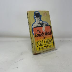 Könnte beinhalten: Ein Vintage-Buch mit dem Titel "The Comedy World of Stan Laurel" von John McCabe. Das Cover zeigt ein Porträt von Stan Laurel mit Melone und einen rot-gelben Titel. Das Buch wird auf einem transparenten Ständer präsentiert.