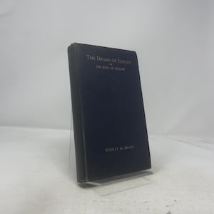 Puede incluir: Un libro de tapa dura azul oscuro titulado "The Drama of Europe or The Soul of History" de Stanley De Brath. El libro tiene letras doradas. El libro está de pie sobre un soporte transparente.