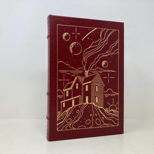 May include: A burgundy hardcover book with a gold illustration of a house with smoke coming from the chimney. The house is on a hill with stars and moons in the sky.