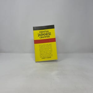 Puede incluir: Un libro amarillo y rojo titulado "ESSENTIAL JAPANESE GRAMMAR" está de pie. La portada del libro presenta el título en letras negras en negrita, con texto adicional debajo. El libro está diseñado para ayudar con el habla y la comprensión.