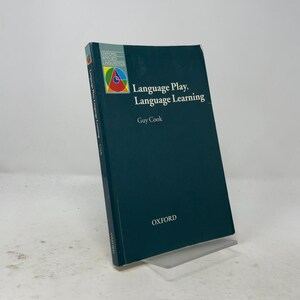 May include: A dark green softcover book titled Language Play, Language Learning by Guy Cook. The cover features a small square graphic with a colorful geometric design in the top left corner and the word OXFORD printed in white at the bottom.