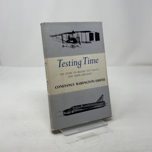 Könnte beinhalten: Ein Vintage-Buch mit dem Titel "Testing Time" mit einem grauen Einband mit Abbildungen von Flugzeugen. Der Titel des Buches und der Name des Autors sind deutlich sichtbar. Das Buch steht auf einem transparenten Ständer.