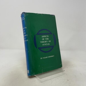 May include: A hardcover book titled Aspects of the Theory of Syntax by Noam Chomsky. The book features a dark green cover with a blue circular graphic design in the center and white text. The spine is blue with white text and a small logo.