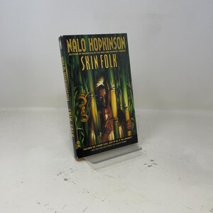 May include: A hardcover book titled "Skin Folk" by Nalo Hopkinson. The cover features a dark-skinned person with a snake, set against a backdrop of green bamboo stalks. The author's name is at the top.