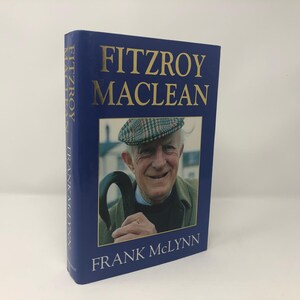 May include: A blue hardcover book with gold lettering titled "Fitzroy Maclean" by Frank McLynn. The cover features a black and white photo of a man wearing a tweed hat and holding a walking stick.