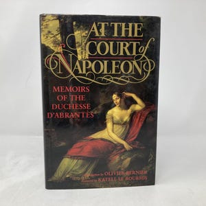 Puede incluir: Un libro de tapa dura titulado "At the Court of Napoleon" con el subtítulo "Memoirs of the Duchesse D'Abrantès". La portada presenta una pintura de una mujer con un vestido blanco y rojo. El libro es una memoria histórica.