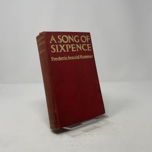 May include: A red hardcover book titled "A Song of Sixpence" by Frederic Arnold Kummer. The title and author's name are in gold lettering. The book is displayed on a clear plastic stand.