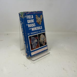 May include: A vintage blue "Field Guide to Rocks and Minerals" book by Frederick H. Pough. The book has a white and blue cover with the title in large white letters. The book is on a clear plastic stand.