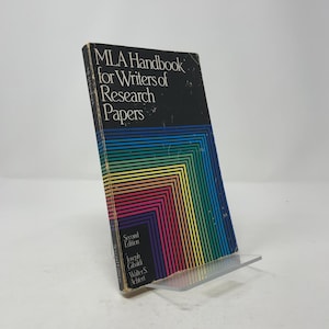 May include: A vintage book titled "MLA Handbook for Writers of Research Papers" in a clear plastic display stand. The cover is black with the title in white and a colorful geometric design. The book is a second edition.