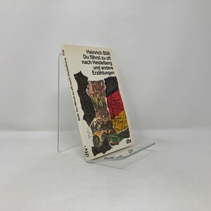 May include: A paperback book titled "Heinrich Böll: Du fährst zu oft nach Heidelberg und andere Erzählungen" is displayed on a clear acrylic stand. The book cover features a collage with a map and text. The book is in German.