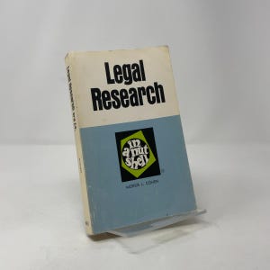 May include: A vintage book titled "Legal Research" with a light blue and white cover. The book features the text "in a nut shell" and the author's name, Morris L. Cohen. The book is displayed on a clear plastic stand.